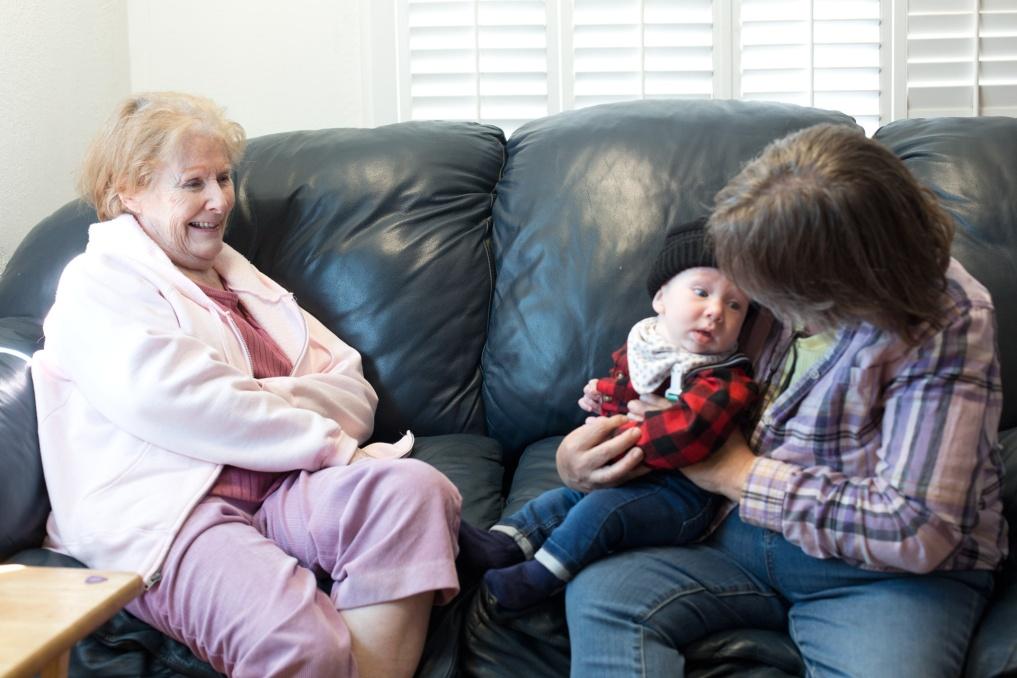 An older woman with light hair, wearing pink, seated on a leather couch and smiling toward a woman holding a baby.